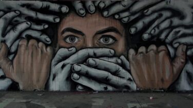 Un’opera di street art realizzata a Berlino raffigura il volto di una persona circondata da tante mani, due delle quali sono posizionate sulla bocca impedendole di parlare